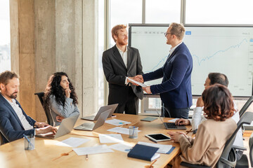 A group of business professionals are gathered around a large conference table. Two men in suits are shaking hands while standing, signaling a successful agreement or partnership