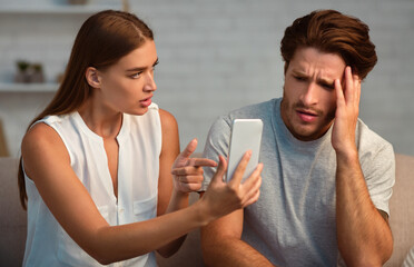 Infidelity Concept. Wife Showing Her Cheating Husband His Cellphone Demanding Explanation Sitting On Couch At Home.