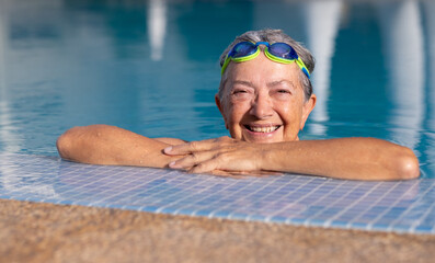 Happy active senior woman doing sport in the outdoor swimming pool looks at camera smiling. Elderly woman and healthy lifestyle in retirement or vacation