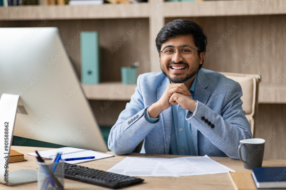 Aufkleber indian man wearing glasses smiles while sitting at his desk, with a computer and papers in front of 