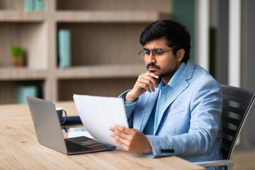 Fototapeta premium A young Indian businessman sits at a desk in an office, reviewing documents while contemplating their contents. A laptop is open on the desk beside him