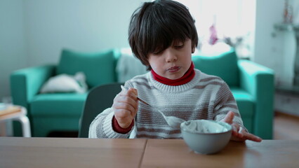 Young boy eats yogurt with a spoon while seated at the table, focusing on each spoonful, capturing...