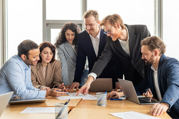 A group of business professionals are gathered around a table in a modern office, collaborating on a project. They are reviewing documents and using laptops.