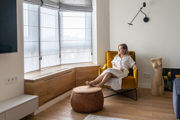 woman in a white dress sits on a chair in a hotel and looks out the window