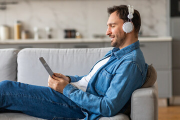 A man wearing headphones sits on a gray couch in a living room, looking at a tablet computer and...