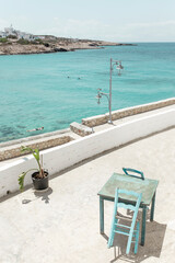 Fototapeta premium A serene spot by the turquoise sea in Donousa features a quaint table and chair beside a vibrant plant, creating an inviting atmosphere for relaxation.