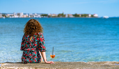 woman sits on coast of adriatic sea with aperitif drink watching panorama of Zadar in Croatia