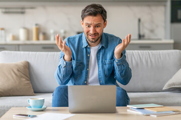 A man sits on a light-colored couch with his hands up in the air looking at a laptop in disbelief....