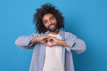 A cheerful curly individual expresses love by forming a heart shape with his hands, standing...