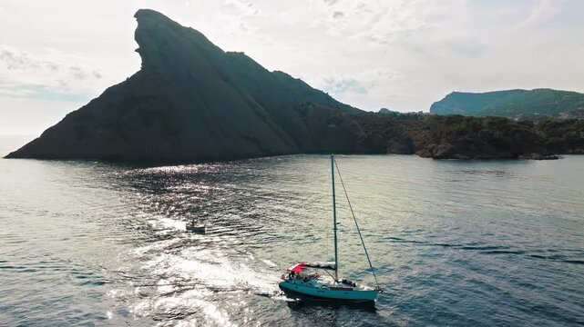Aerial view of a boat leaving a Botanical garden with Snug Cove framed by rugged cliffs in Parc du Mugel. View from above of ship departing a pebble beach with turquoise clear water in France.