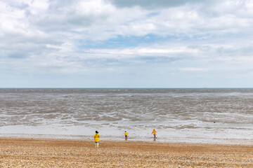 Obraz premium Woman and two children in bright yellow rain gear on pebblestone beach and coastline with mudflats and horizon near Littlestone-on-Sea, Kent, UK with white clouded sky