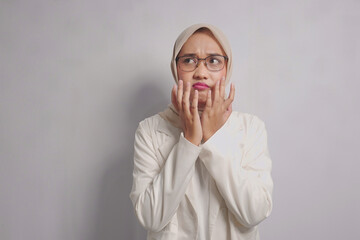 Woman in a white blazer and hijab, looking concerned and holding her hands to her face.