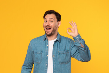 A man with brown hair and a beard, wearing a blue denim shirt over a white t-shirt, smiles while showing the okay gesture with his right hand, standing against a yellow background.