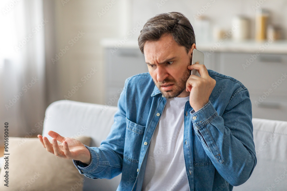 Poster A man sits on a couch in a living room, frowning and gesturing with his hand while talking on a phone. He is frustrated and angry, and it seems like he is in the middle of a heated conversation.