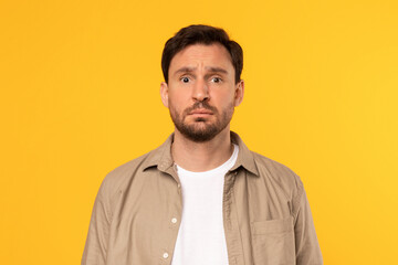 A man with a beard and brown hair is wearing a beige button-up shirt over a white t-shirt. He is looking directly at the camera with a serious expression on his face. The background is a solid yellow.