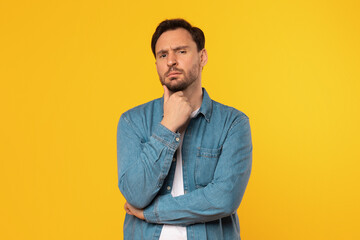 Man with a beard, wearing a blue denim shirt, stands in front of a yellow background, looking directly at the camera with a thoughtful expression. He has his hand on his chin and his arms are crossed