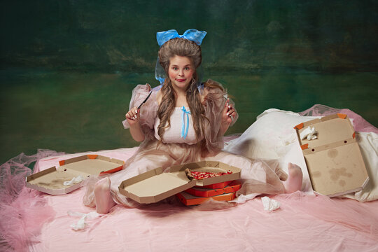 Sleepover. Elegant young woman nystatin in bed with many boxes and eating pizza with positive face against vintage green background. Bedroom party. Concept of history, baroque art, comparison of eras