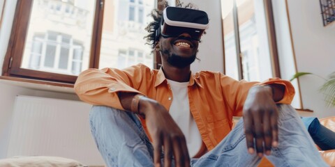 Fototapeta premium A man in an orange shirt is sitting on a couch with his hands on his knees. He is wearing a virtual reality headset and he is smiling