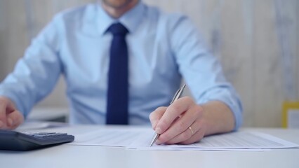 Unknown man accountant with blue t-shirt concentrating while calculating costs and taking notes at his desk. Taxes, audit in business