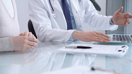 Doctor is consulting patient in modern fair clinic. Healthcare professional discusses medical information with a woman, pointing into the laptop computer monitor. Medicine concept