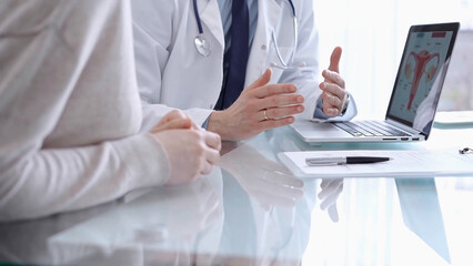 Doctor is consulting patient in modern fair clinic. Healthcare professional discusses medical information with a woman, pointing into the laptop computer monitor. Medicine concept