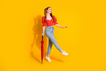 Full body length photo of young lady in red blouse and jeans holding umbrella strolling with parasol isolated on yellow color background