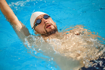 Swimmer in white cap and goggles powers through water, maintaining steady freestyle stroke as he trains in brightly lit outdoor pool. Concept of sport, competition, active and healthy lifestyle. Ad