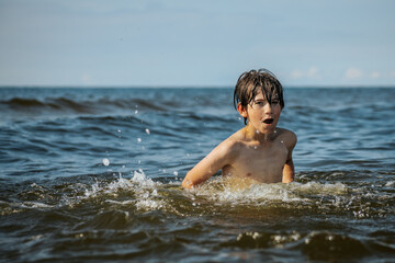 A boy joyfully splashes in the water with arms raised, capturing the excitement of playing in the ocean on a sunny day. The scene embodies the carefree spirit of childhood and summer fun