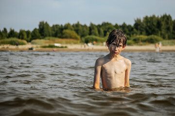 Fototapeta premium A smiling boy with arms outstretched stands in the water at a beach. Other beachgoers are visible in the background under a clear sky