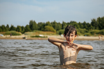 A smiling boy with arms outstretched stands in the water at a beach. Other beachgoers are visible in the background under a clear sky