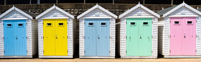 A row of five white beach huts on the promenade at Lyme Regis, west Dorset, on the south coast of England. Doors painted in pastel shades of blue, yellow, green and pink.