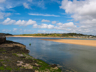 River Camel estuary between Padstow and Rock in Cornwall, England. Seaweed covers foreground shore. Shows sandbank and grassy hills in background. Cloudy blue sky. 
