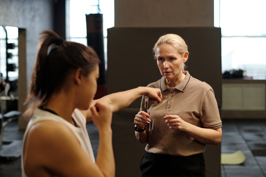 Blond mature woman in activewear standing in front of young female boxer practicing kicks and giving her advice about techniques - Powered by Adobe