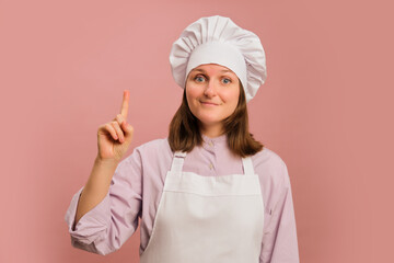 Woman chef pointing finger up on studio pink background. Portrait of a female person in chef's clothing