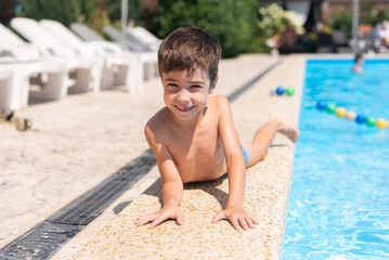 A little boy lies on his stomach on the edge of the open air pool and smiles at the camera