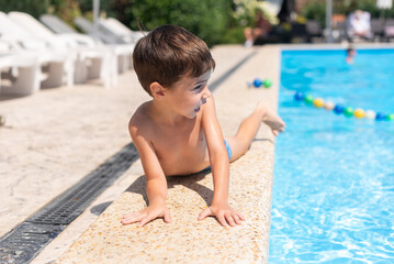 A little boy lies on his stomach on the edge of the open air pool and smiles at the camera