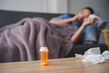 Prescription medicine bottle with pills on a wooden coffee table and a sick person resting on a couch in the background