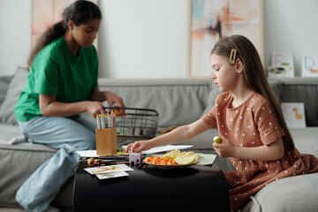 Side view of cute little girl in casual apparel taking crayon and having piece of fresh apple while sitting by table against teenage female sitter