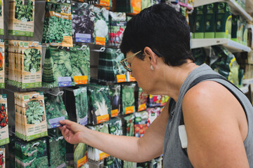 A mature woman selects packaged vegetable seeds from a shelf in front of a display case of samples...