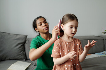 Cute little girl in casual dress counting and bending her fingers while standing in front of baby sitter brushing child hair with hairbrush