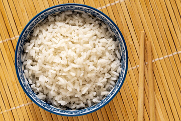 Cooked white rice in an asian white and blue ceramic bowl next to chopsticks isolated on light bamboo matt from above.
