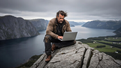 Ruggedly independent freelance writer perched on jagged outcropping above serene Scandinavian fjord and countryside. Immersed in work, natural backdrop