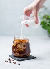 Iced coffee latte in a glass with a hand pouring milk on a light  background with coffee beans and branch.