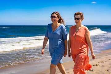 Summer vacation. Two pretty mid-adult women wearing flowing dresses and sunglasses walking on the...