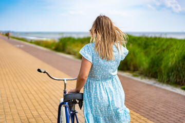 Summertime activity. Woman with long blond hair wearing blue dress walking with blue cruiser bike on bicycle path on seafront on sunny day. Back view.