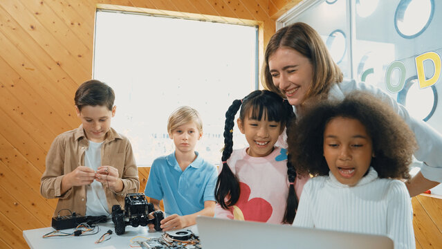 Caucasian teacher praise student while looking at learner work or presentation. Group of diverse student looking at presentation and fixing motherboard at table with chips and wires placed. Erudition.
