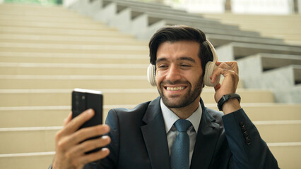 Happy smart business man taking selfie while smiling at smart phone. Closeup image of professional executive manager sitting at stairs while wearing suit and headphone. Creative business. Exultant.