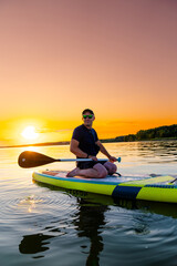 Man Paddleboarding at Sunset. A man kneels on a paddleboard, holding a paddle in front of him as he looks at the camera, with the setting sun behind him.