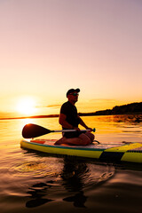 Man Paddleboarding at Sunset. A man kneels on a paddleboard as he paddles across a still lake at...