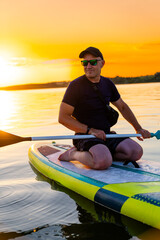 Man Paddleboarding at Sunset. A man kneels on a paddleboard while paddling across a calm lake at...